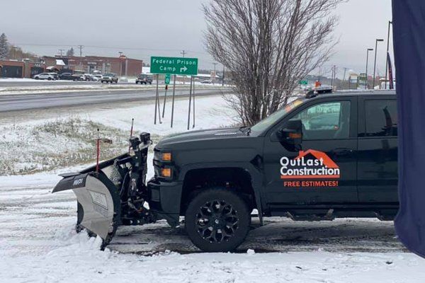 Black snowplow truck clearing snow on a snowy roadside near a highway sign.