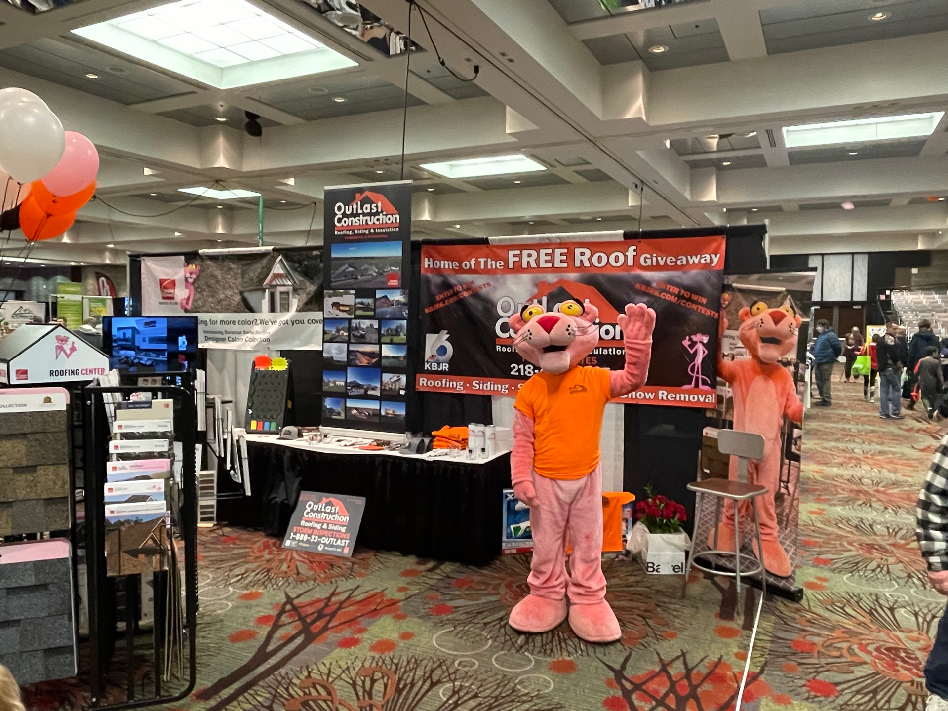 A pink panther mascot is standing in front of a booth at a convention.