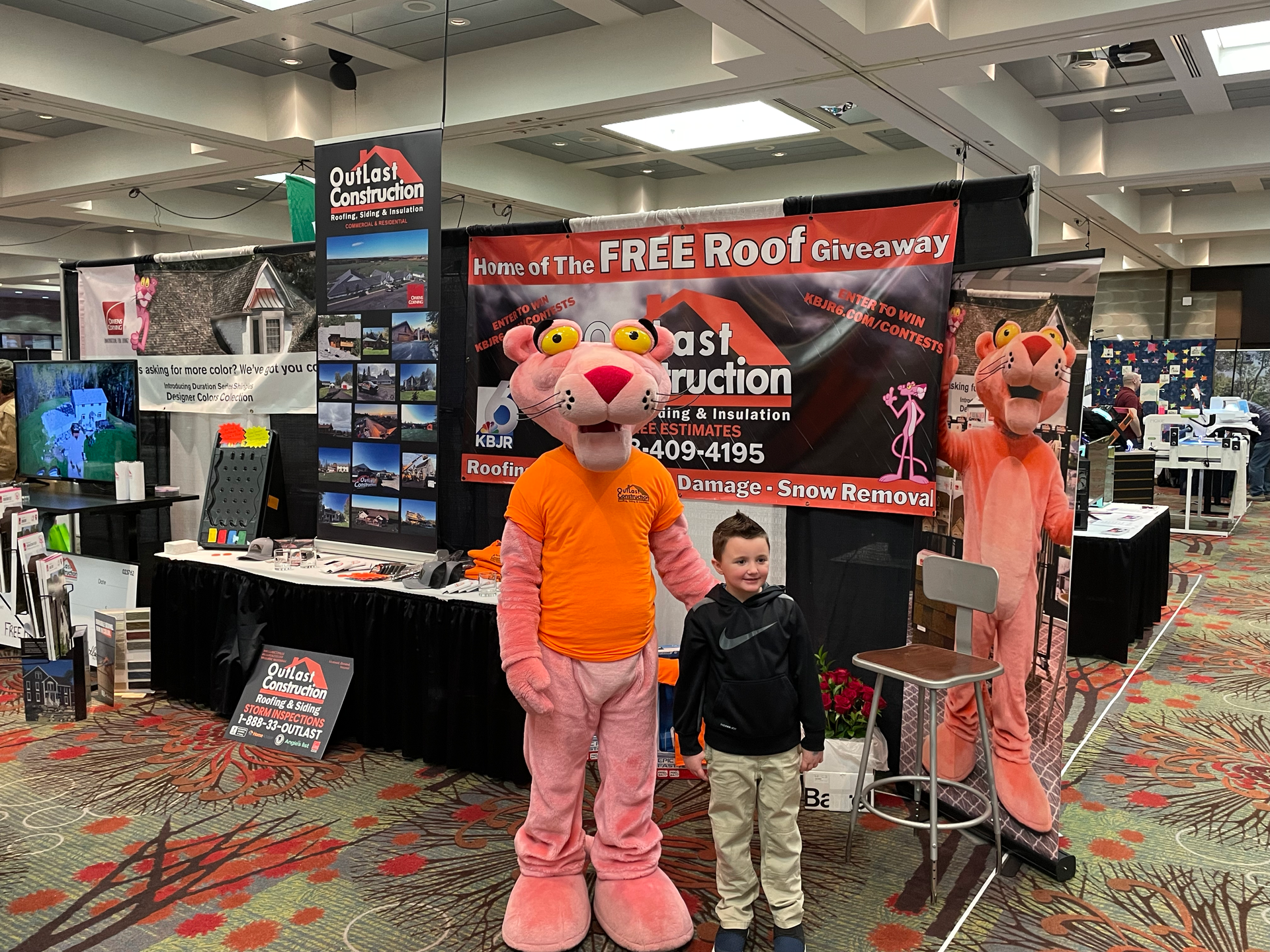 A boy is standing next to a pink panther mascot at a roofing convention.