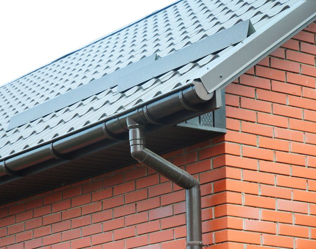 A brown metal gutter and downspout attached to the edge of a roof with dark gray shingles above a red brick wall.