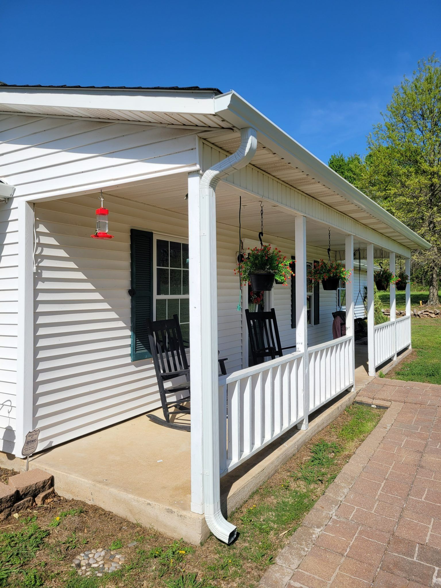 A white house with a covered porch featuring two black rocking chairs, hanging flower baskets, and a brick walkway.