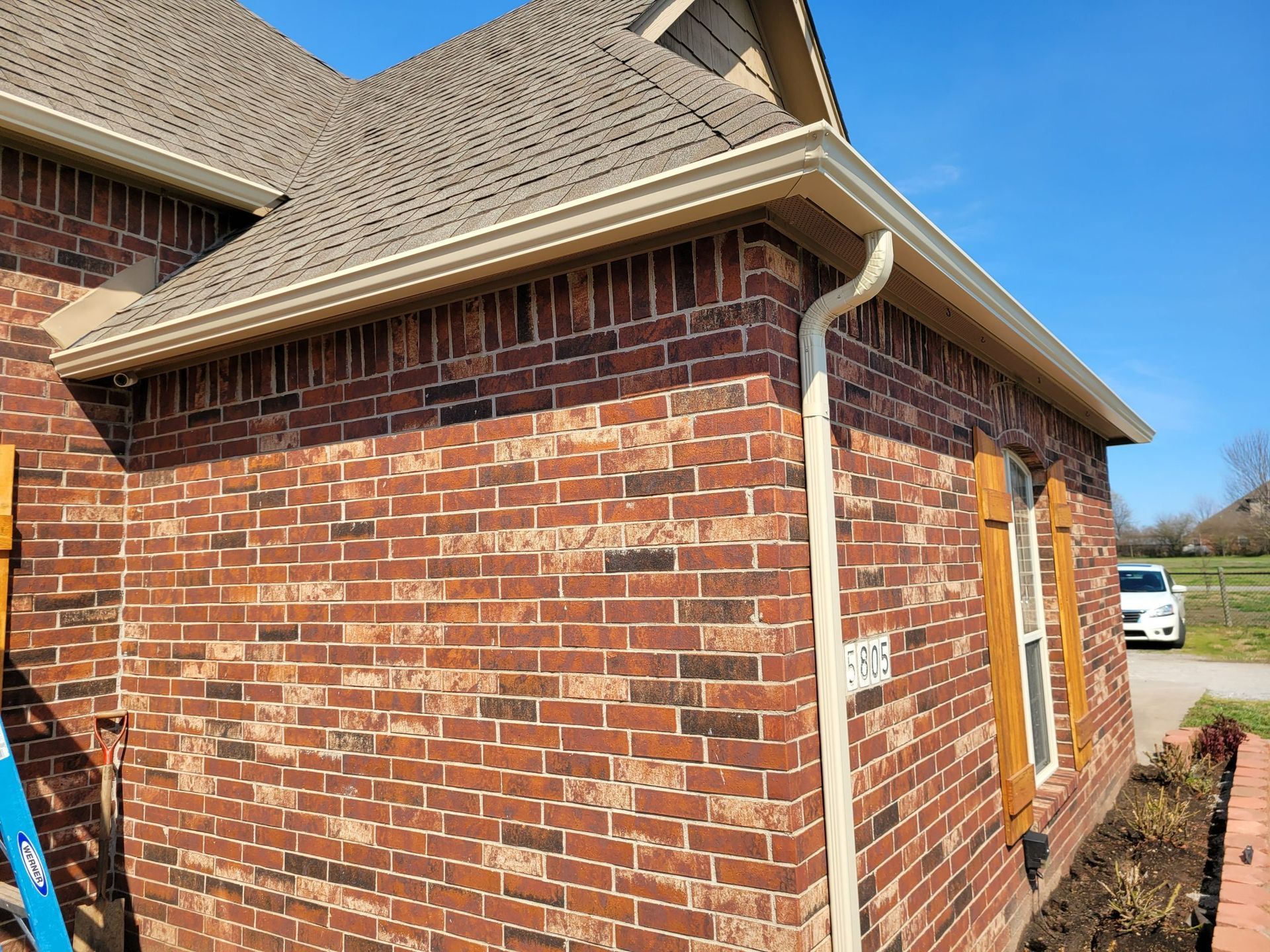 A red brick house exterior with a tan gutter, downspout, and window shutters, under a clear blue sky.