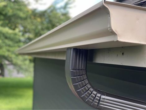 A tan-colored metal rain gutter attached to the edge of a roof with a dark gray corrugated downspout elbow underneath.
