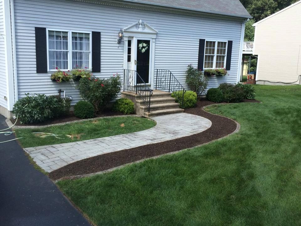 A house with a brick walkway leading to the front door. Landscaped with bushes, flowers and black shutters.