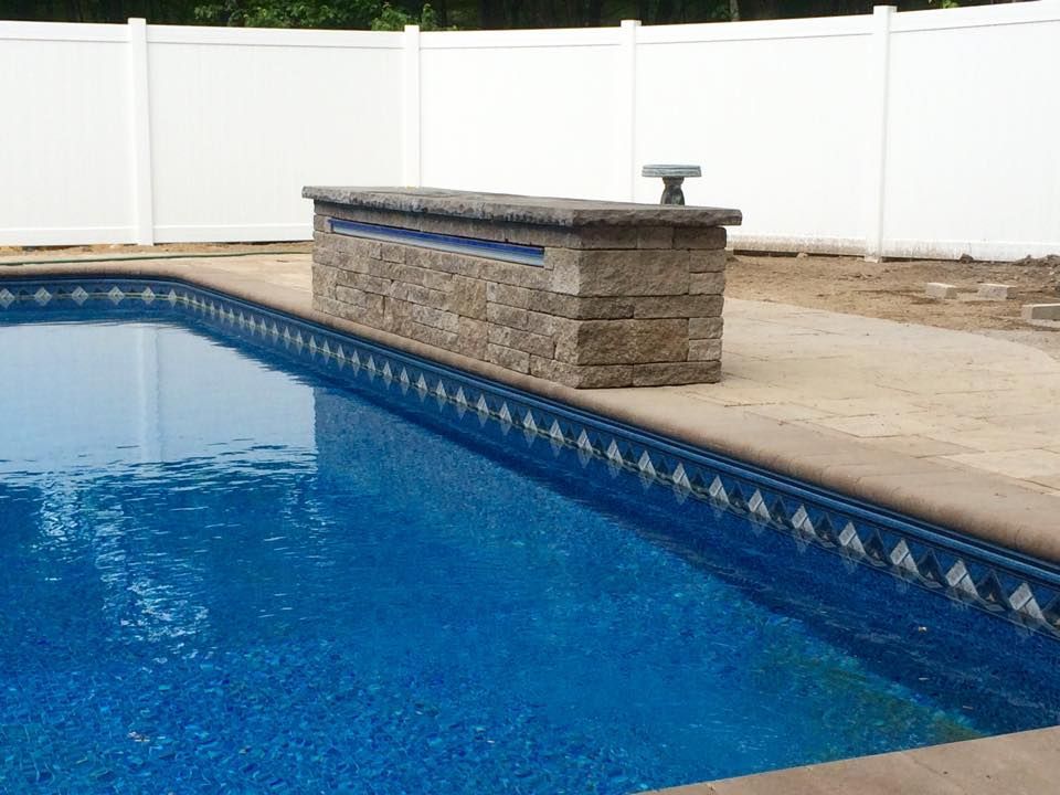 Pool with blue water and stone wall, bordered by a decorative tile. White fence in the background.