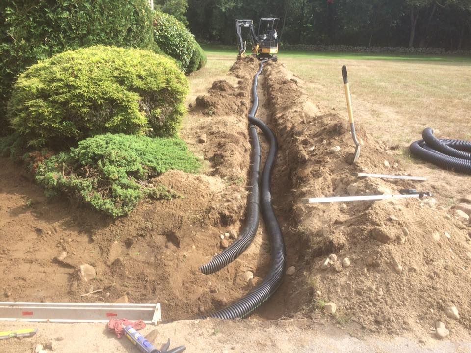 Ditch digging for landscaping, with black corrugated pipes in a trench next to bushes and grass.