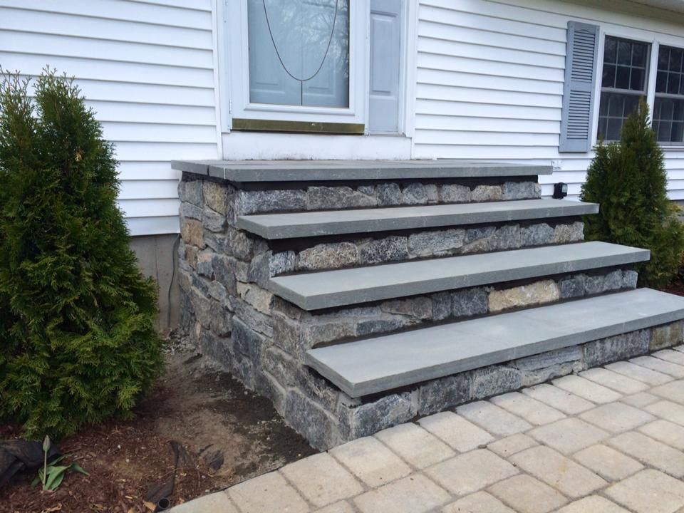 Stone steps leading up to a white house with a blue door. Steps are gray and the side is stone.