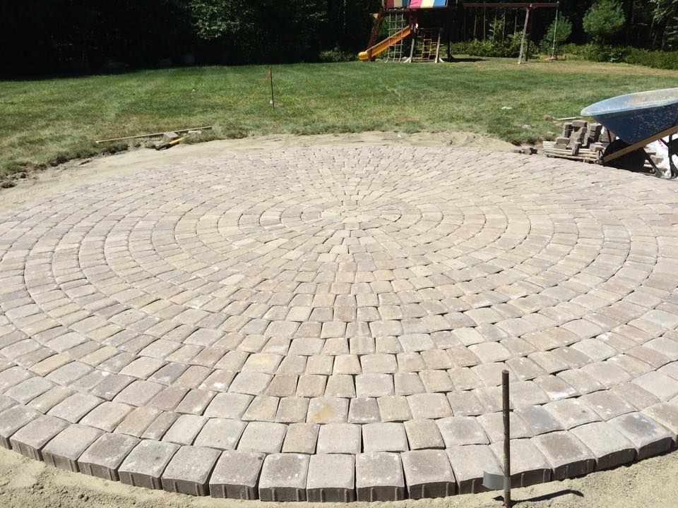 Circular brick patio under construction, set in a green yard, with a playset in the background.