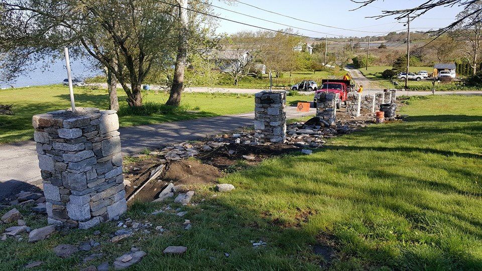 Stone pillars partially constructed on a grassy area, leading to a road and buildings in the background.