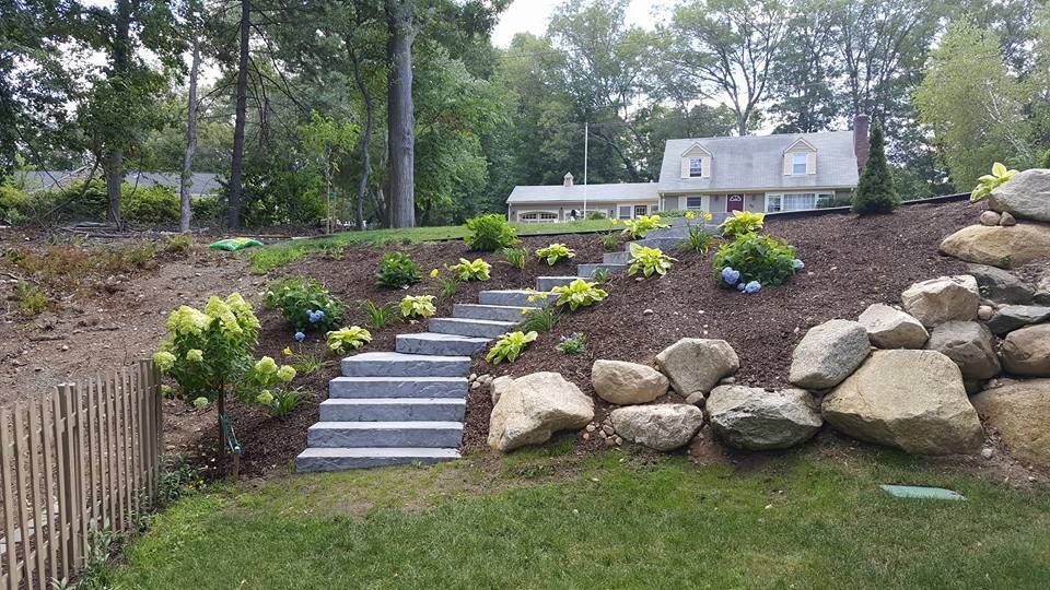 Stone steps and landscaping ascend a hillside towards a house, with rocks and greenery on either side.