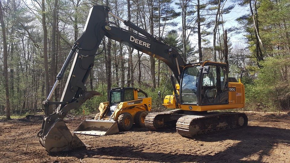 Yellow and black Deere excavator and skid steer on a dirt lot surrounded by trees.