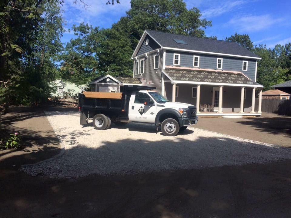 A dump truck on a gravel driveway in front of a two-story house with a porch.