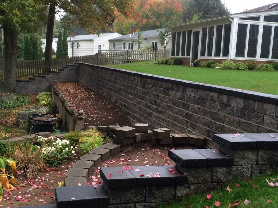 Stone retaining wall with steps, garden, lawn, and houses in the background. Fall foliage.