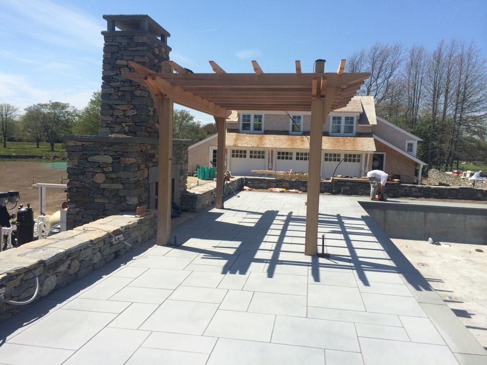 Patio with stone fireplace and wooden pergola; house in background, person working.