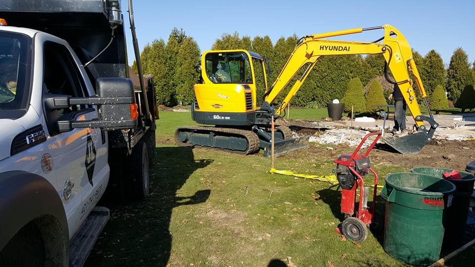 A yellow excavator working near a truck and trash cans in a grassy area.