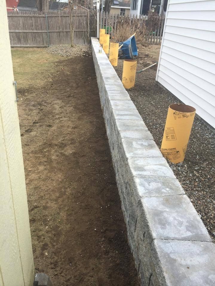 A long, gray retaining wall in a yard, with yellow cylinders and bare earth.