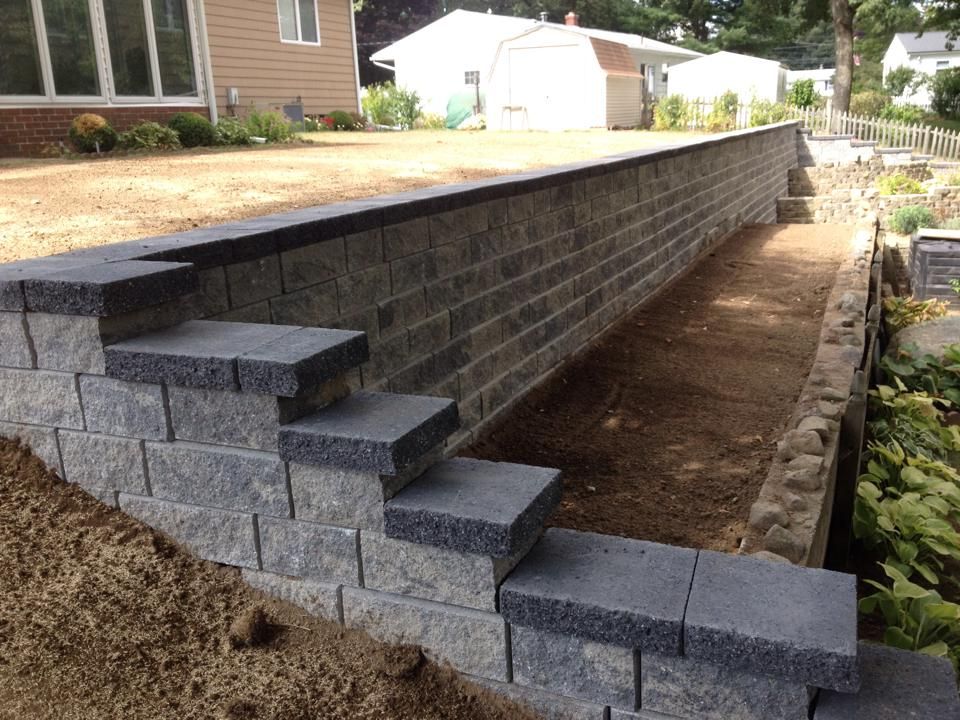 Stone retaining wall with steps leading up to a raised yard, near a house and greenery.