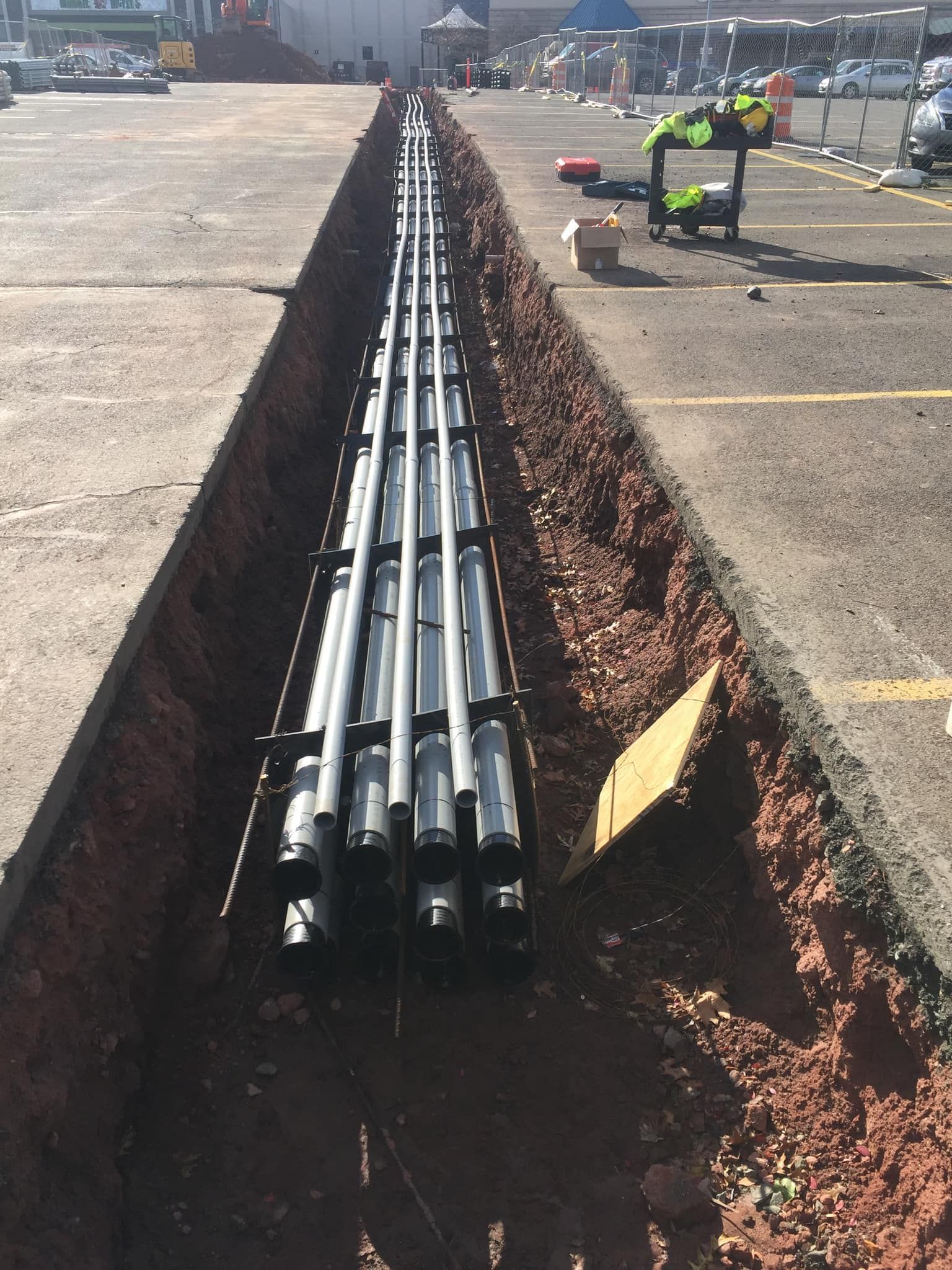 Trench in asphalt with black pipes laid inside; worker in vest near.