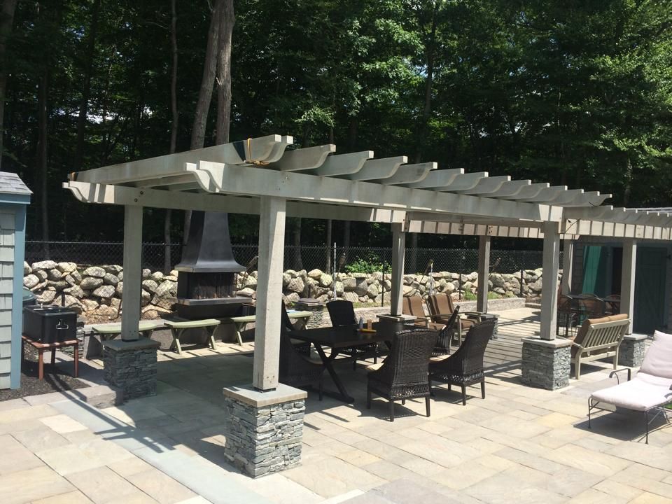 Pergola with dining table and fireplace on patio. Stone columns and backdrop.