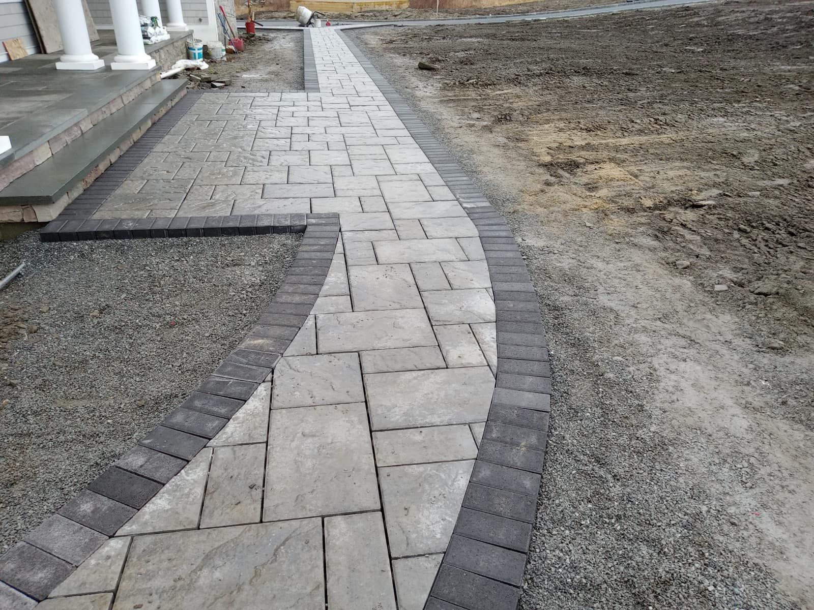 Stone pathway leading from a porch with white columns, edged with dark bricks, in a yard.