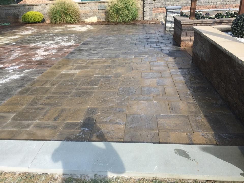 Stone patio with brick wall and landscaping. Brown pavers lead to a concrete walkway.