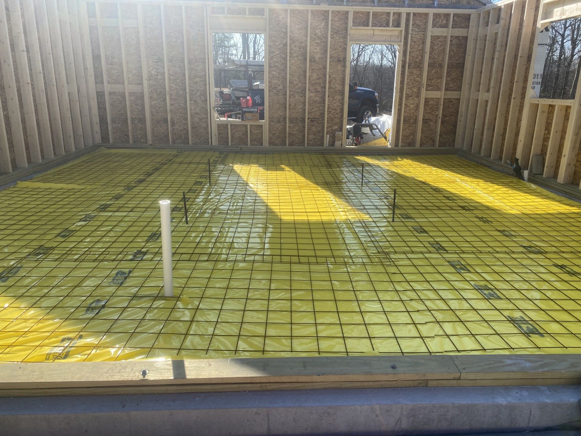 Construction site: framed walls, yellow vapor barrier, rebar grid, white pipe, sunny outdoor setting.