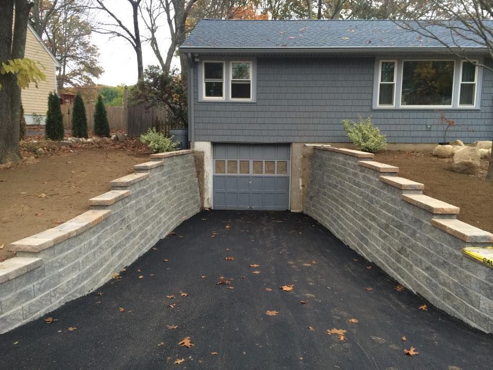 Asphalt driveway between gray retaining walls leading to a garage door under a gray house.