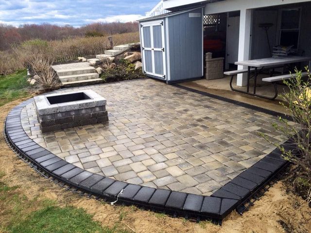 Patio with a fire pit, bordered with dark pavers, next to a shed and picnic table.