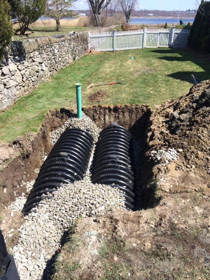 Two black corrugated pipes installed in a trench, surrounded by gravel, green grass, and a green pipe sticking up.