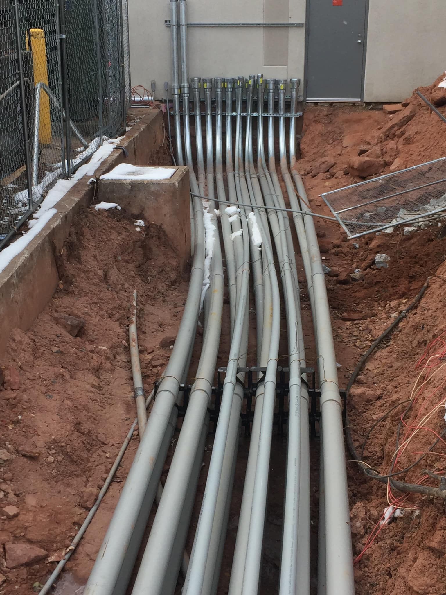 Gray conduits running into a building from a trench. Dirt, snow, and fencing surround the conduits.