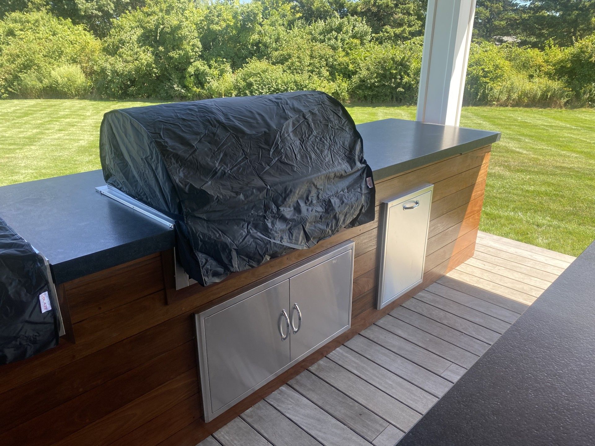 Outdoor kitchen with covered grill, stainless steel doors, and countertop on wooden structure.