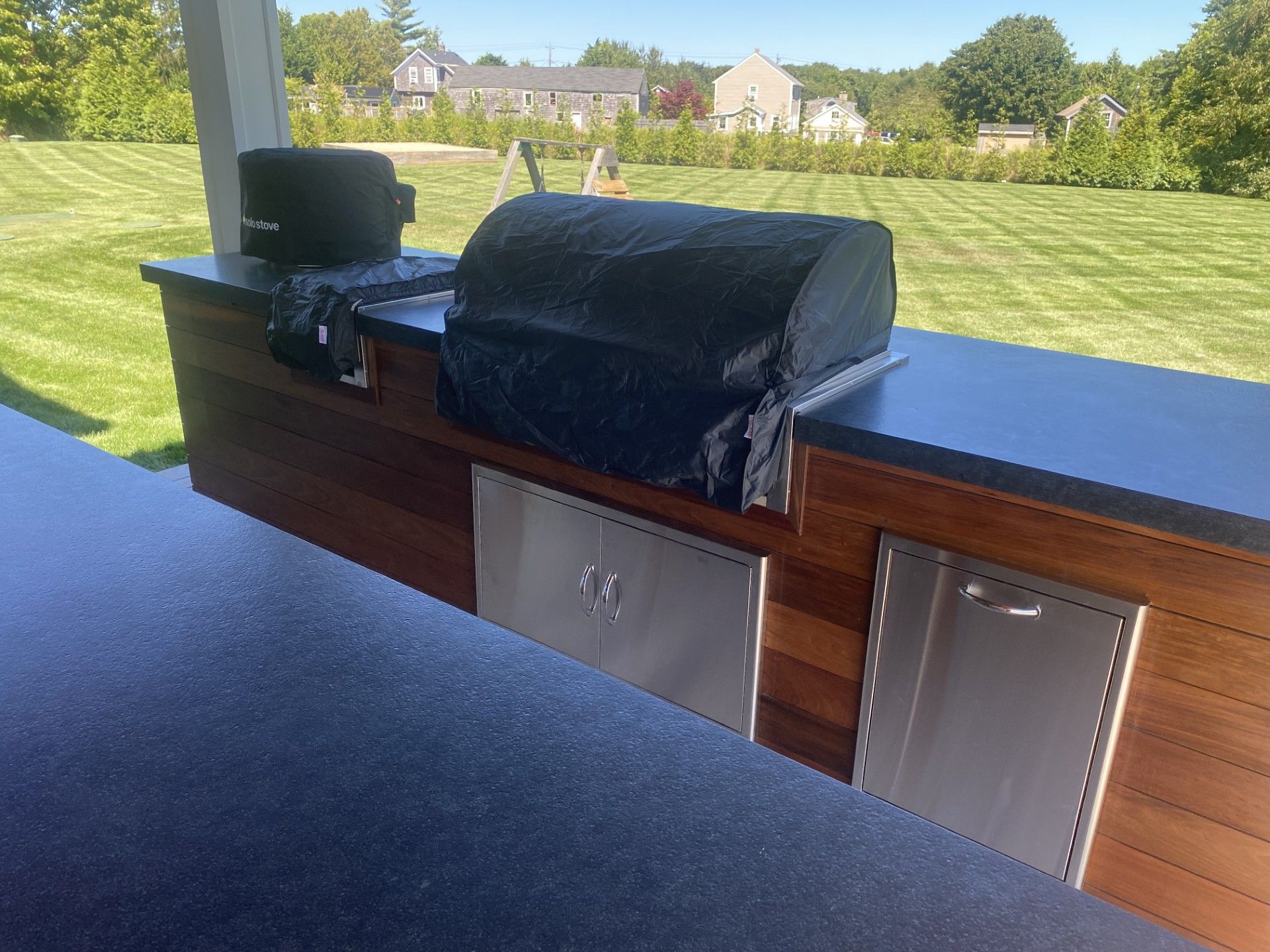 Outdoor kitchen with grill, covered appliances, and stainless steel doors. Wooden cabinets with a lawn in the background.