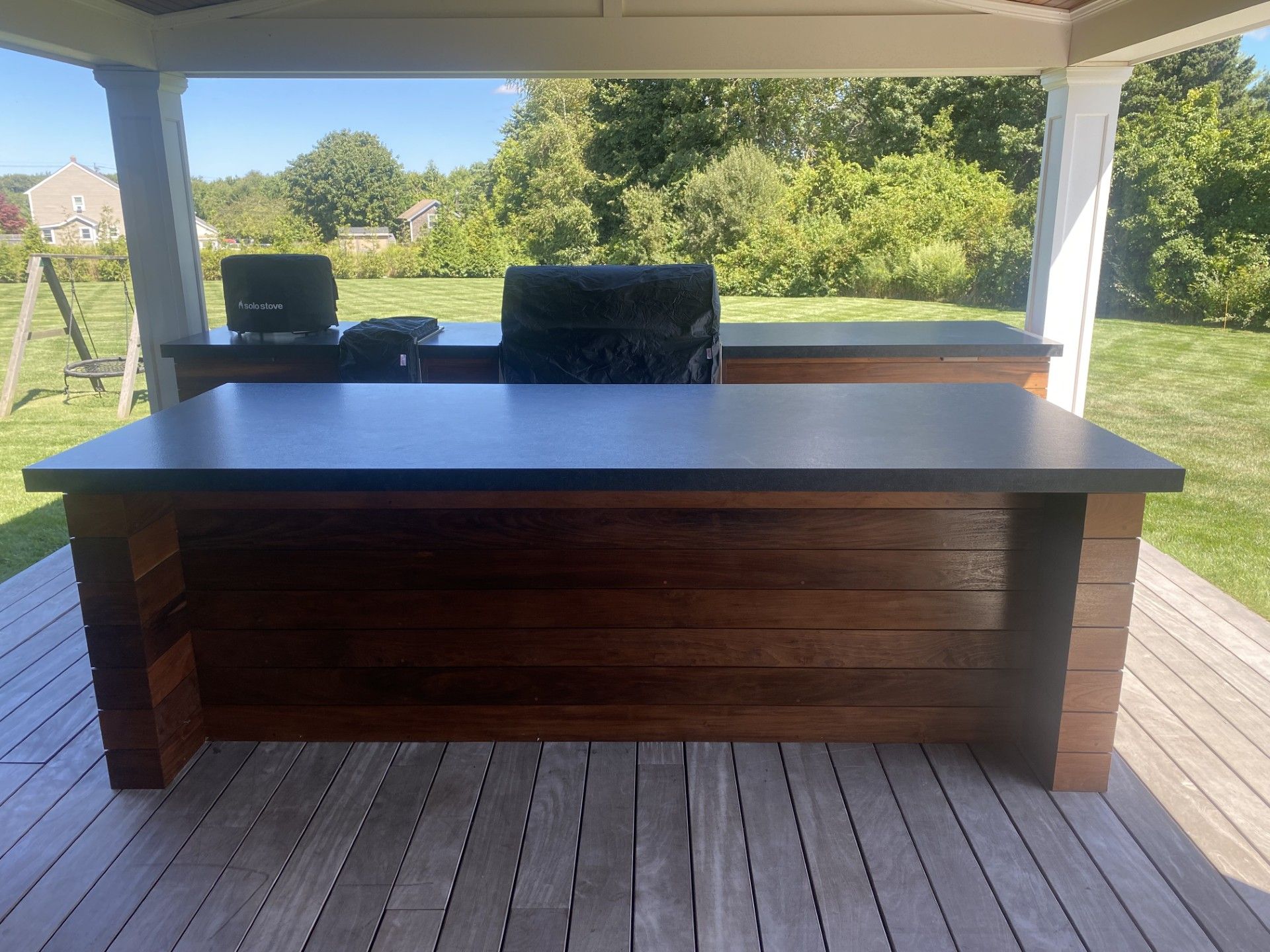 Outdoor kitchen with dark countertop and wood siding under a white-pillared porch.