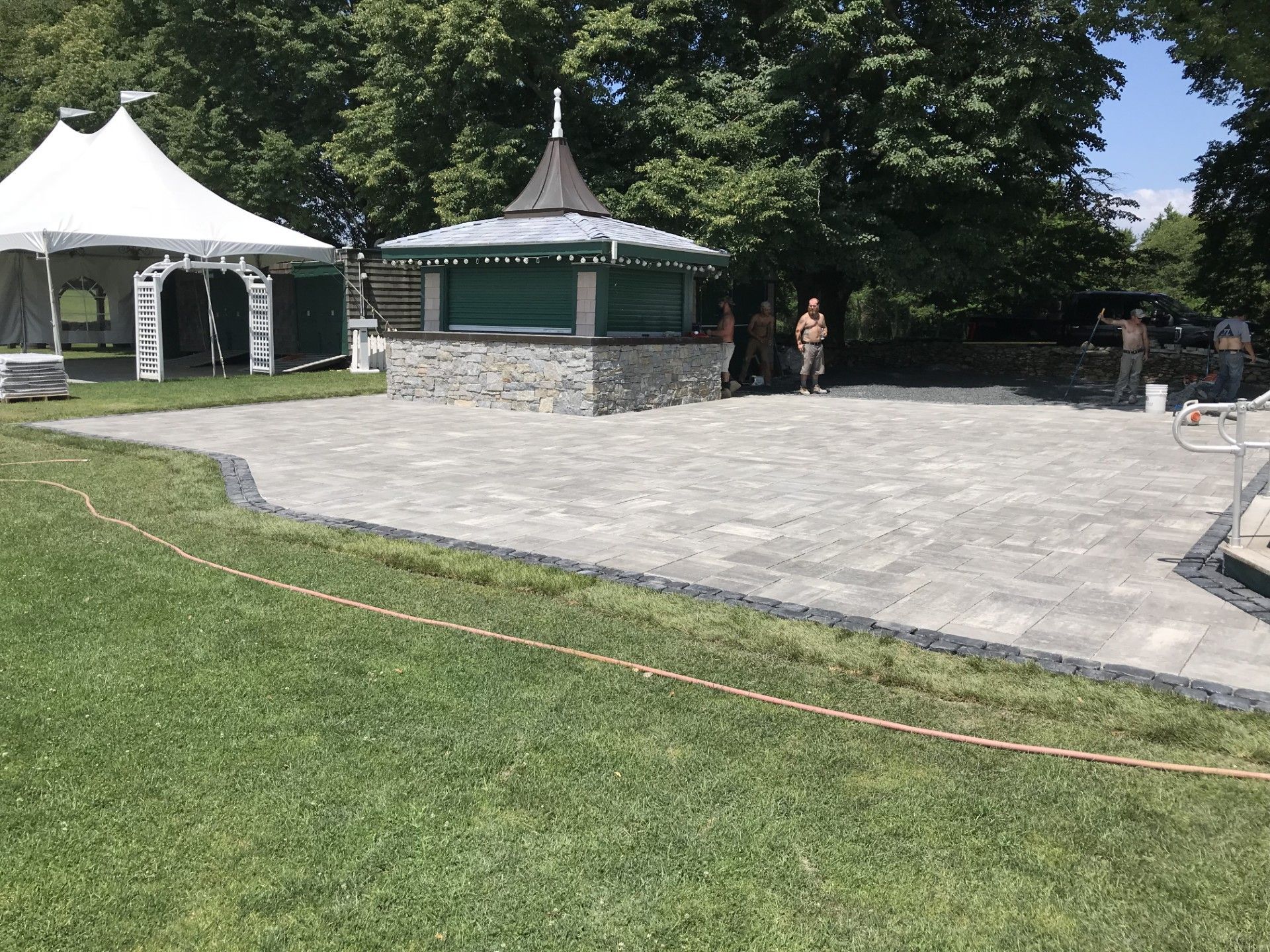 Outdoor paved area with a small stone building and a tent on a grassy lawn. Several people are visible.