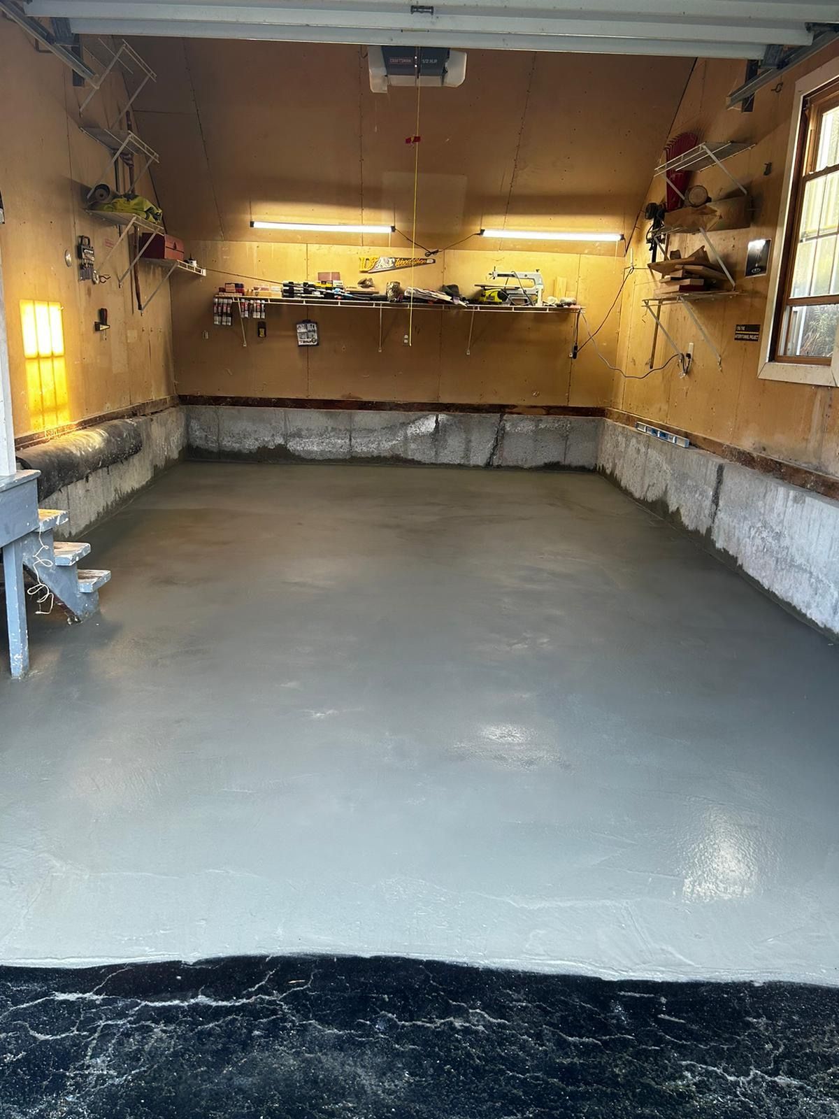 Newly poured grey concrete garage floor. Walls are tan with shelves and tools.