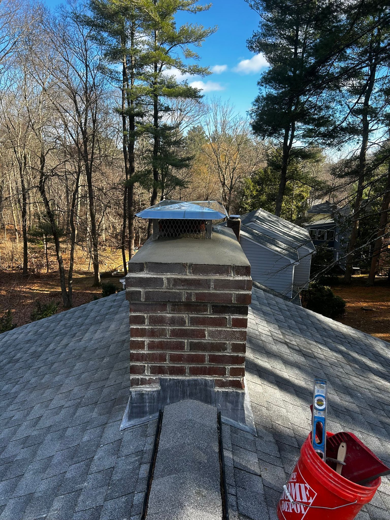 Brick chimney on a gray shingled roof with a chimney cap, set against a forest and blue sky.