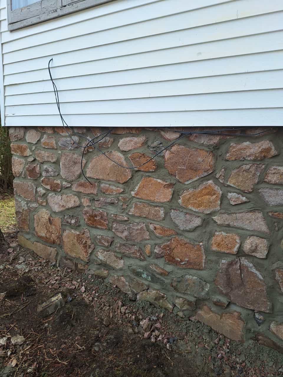 Stone foundation of a house, below white siding. Brown stones set in gray mortar.