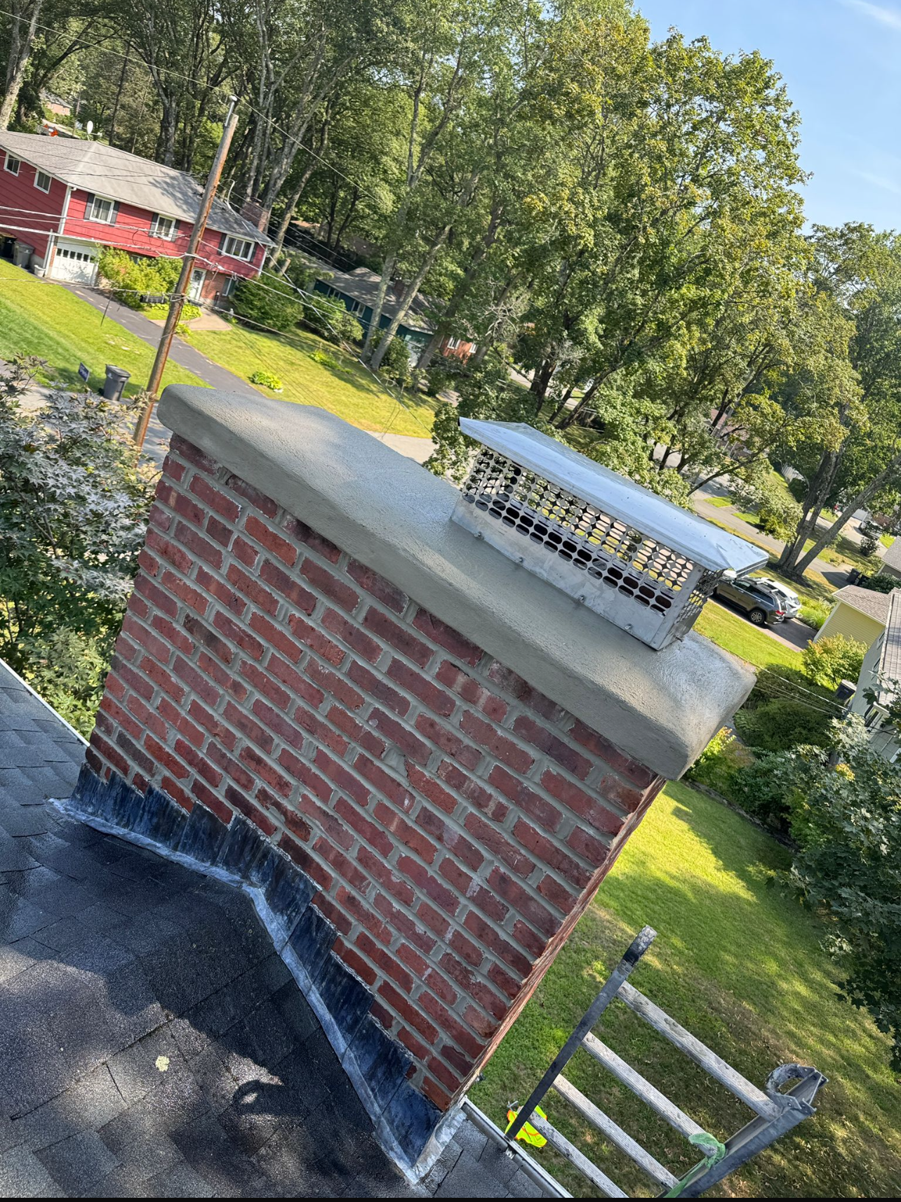 Brick chimney on a black roof, surrounded by green trees and grass, topped with a metal cap.