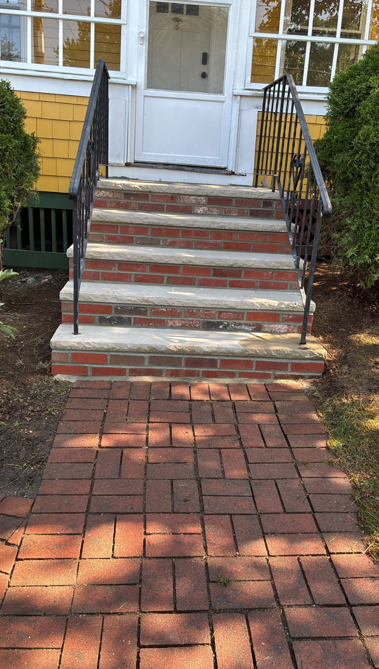 Brick steps leading to a white door, with a brick pathway. Black railing, yellow house.
