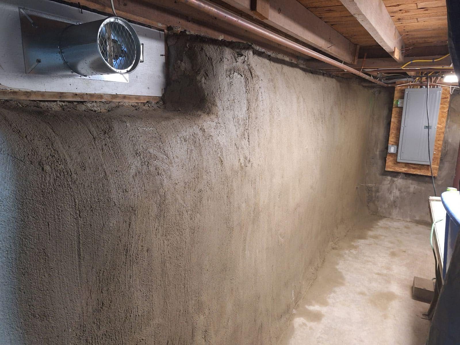 Gray concrete basement wall, partially damp, with overhead ductwork and electrical panel.