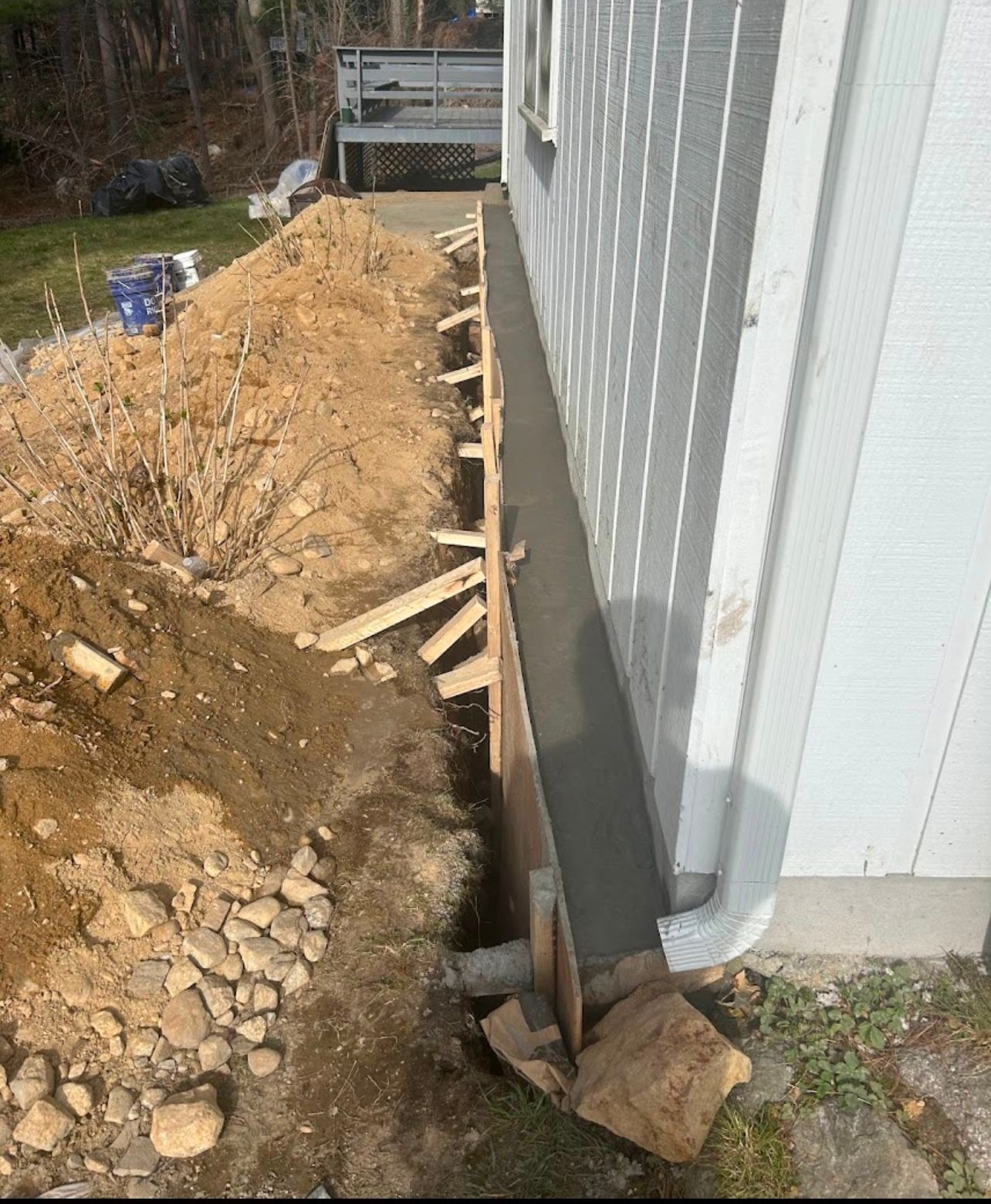 Construction site: concrete foundation poured alongside a white building, with wooden forms visible and earth surrounding.