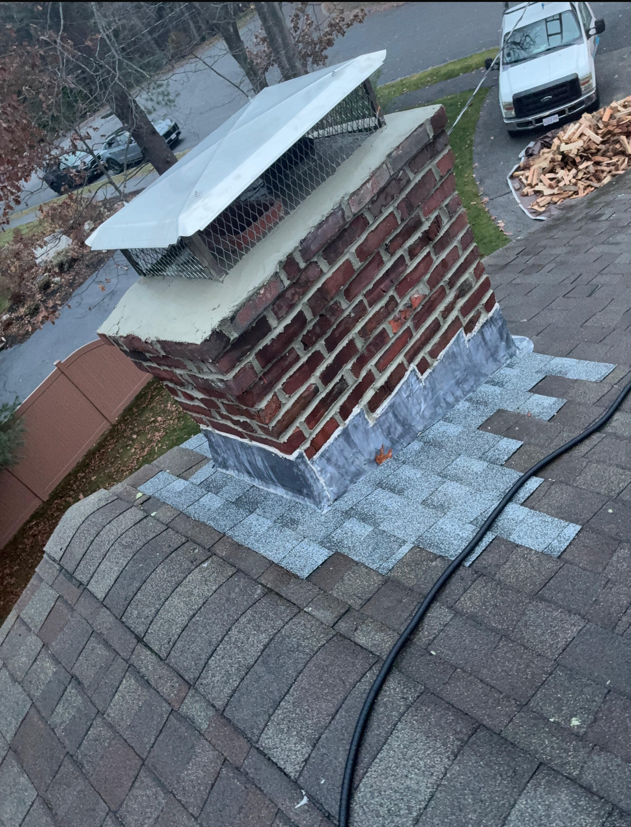Brick chimney on a shingled roof with a metal cap and flashing; a van is parked nearby.