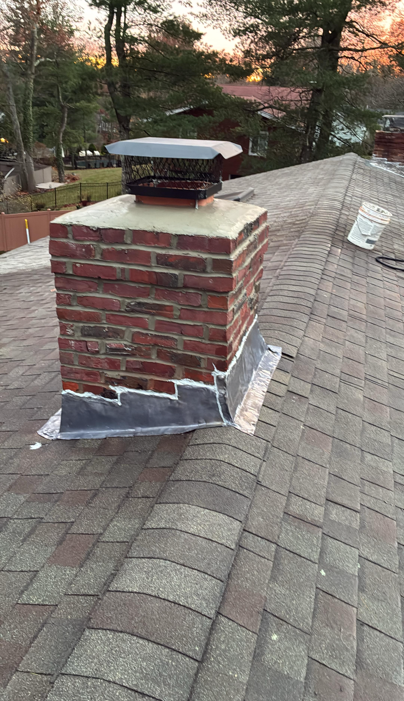 Brick chimney on a shingled roof with a chimney cap, gray flashing, and a backdrop of trees.