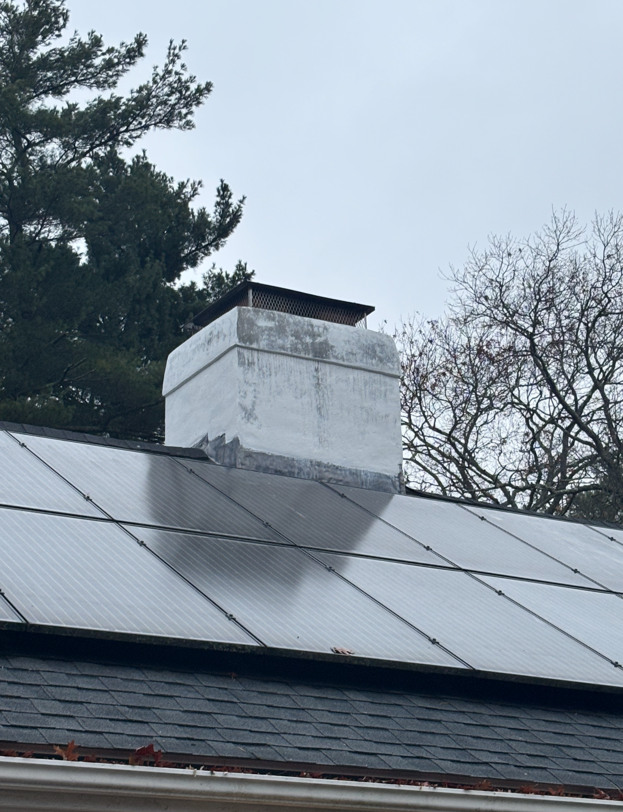Chimney on a roof with solar panels. Gray sky, white-painted chimney, metal cap.