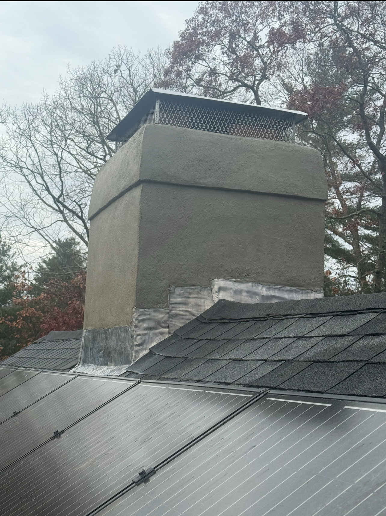 Chimney on a roof with solar panels. Gray stucco, wire mesh top, against a cloudy sky.