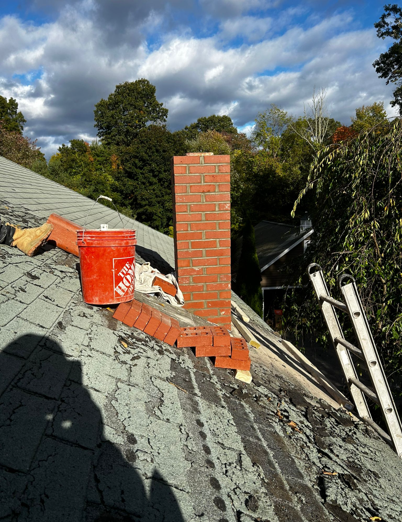 Chimney repair on a roof; bricks, orange bucket, ladder, and trees under a cloudy sky.