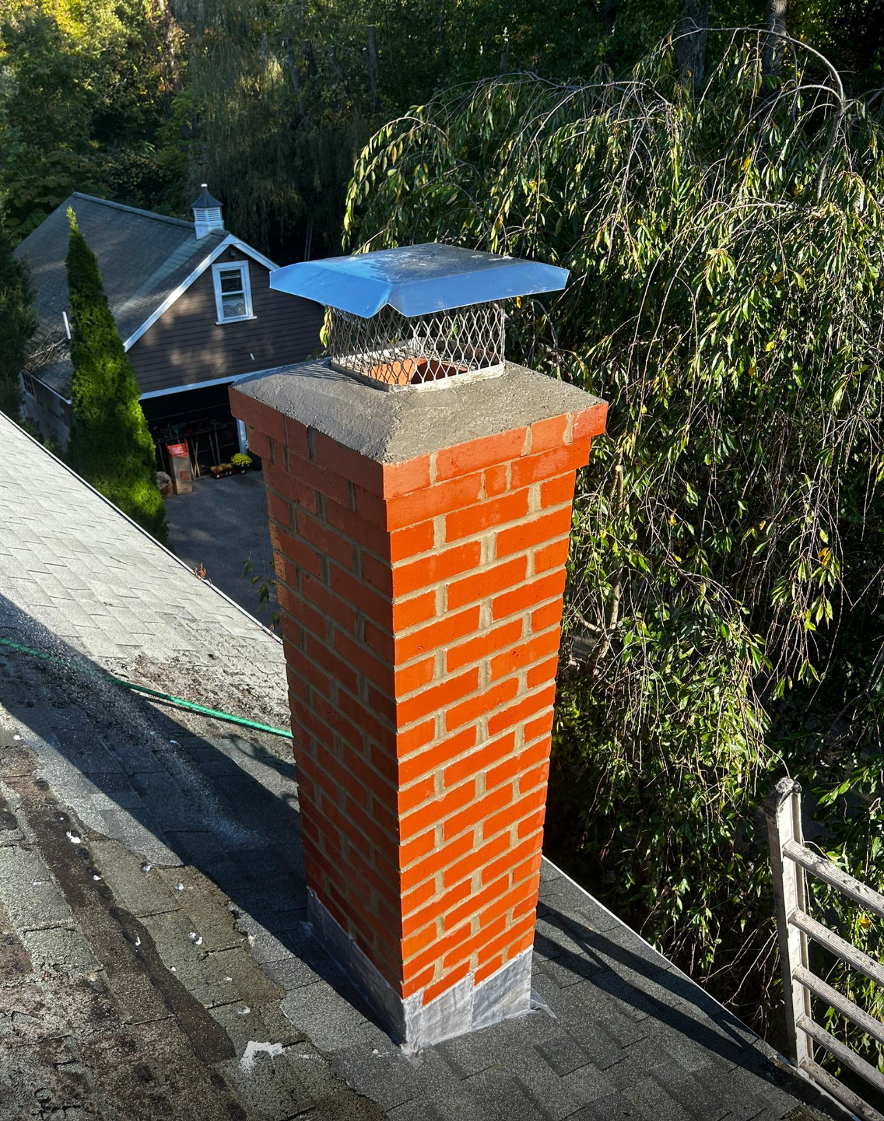 Brick chimney on a roof with a metal cap, background includes a house and trees.