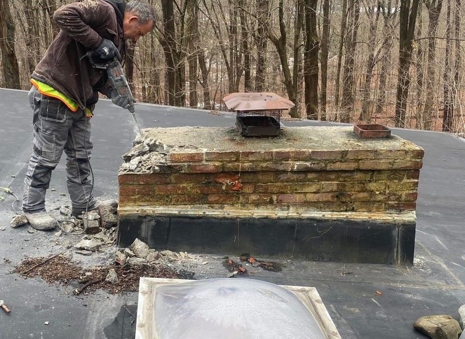 Person demolishing a brick chimney on a rooftop with trees in the background.