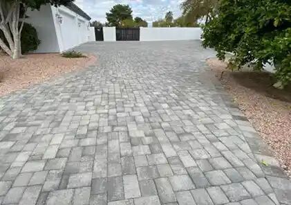 Brick driveway leading to a black gate, bordered by gravel and a white wall.
