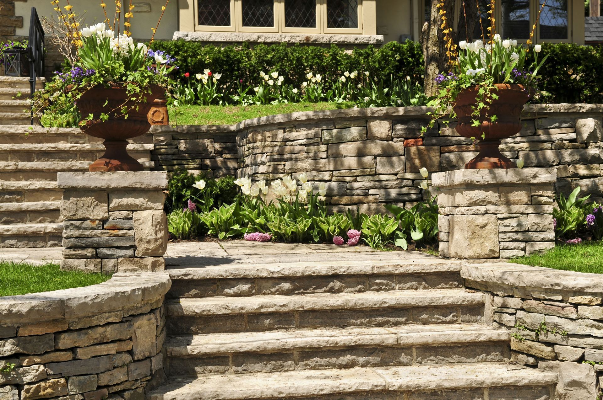 Stone steps leading to a house with planters filled with flowers; grassy area and flowering shrubs.